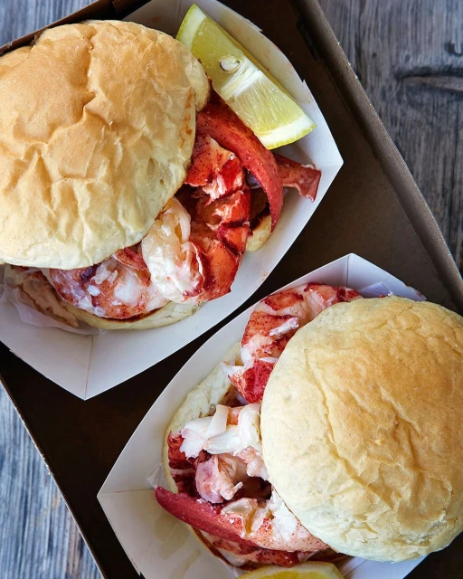Two lobster rolls in paper trays, each topped with a lemon wedge, presented on a wooden surface, capturing a fresh seafood meal.