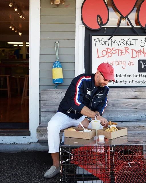 Adult man in a jacket and baseball cap sits outside a seafood restaurant and enjoys takeout. Lobster traps are used as a table.