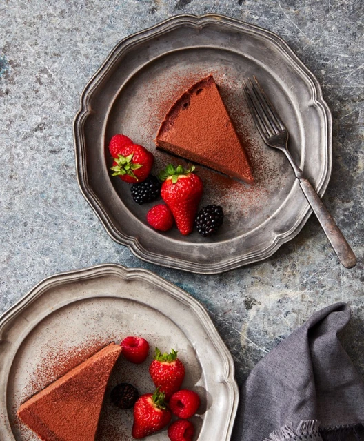 Two antique pewter plates featuring slices of chocolate cake dusted with cocoa powder and garnished with raspberries, strawberries, and blackberries.