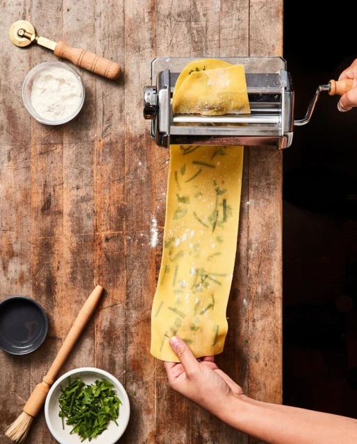 Person using a pasta machine to make pasta with herbs on a wooden surface, surrounded by a pasta cutter, flour, and herbs.