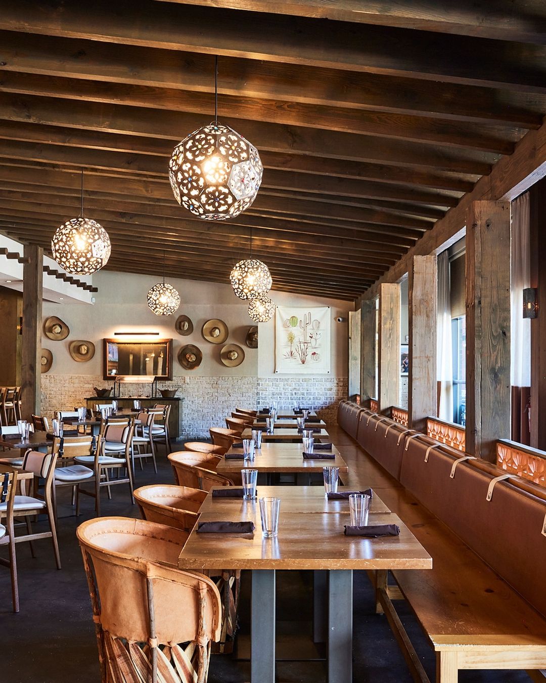 Interior of a restaurant features rows of dining tables and chairs, overhead decorative lighting, and rustic wood beam ceiling.