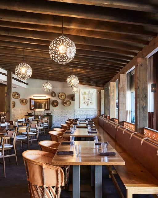 Interior of a restaurant features rows of dining tables and chairs, overhead decorative lighting, and rustic wood beam ceiling.