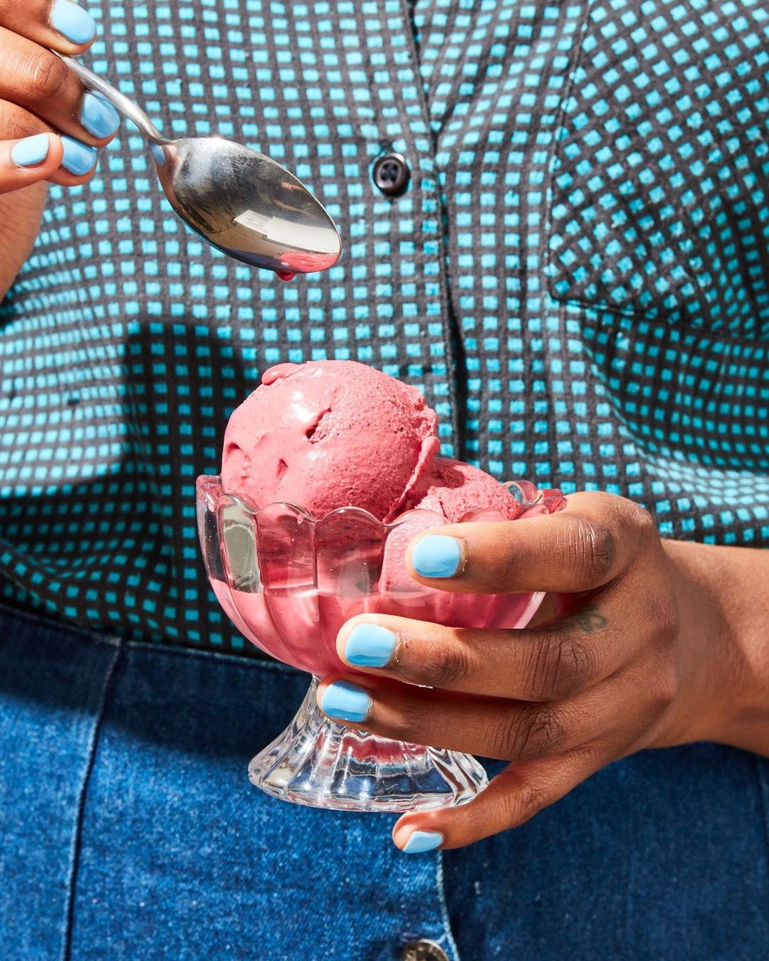 Person holding a glass bowl of pink ice cream topped with a dripping spoon in front of a black-and-turquoise patterned shirt.