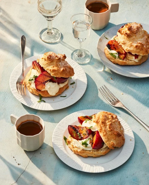 Overhead shot of three plum scones with cream on plates with forks, coffee mugs, and stemmed glasses on a light blue table.