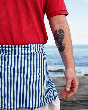 Man wearing red polo shirt and blue-and-white striped apron with lobster tattoo on arm by ocean