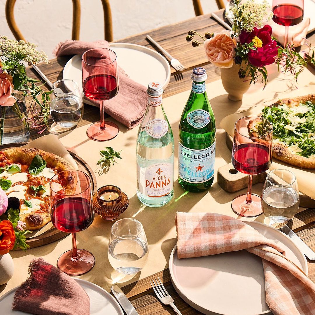 A pizza and wine meal setup on a wooden table with water bottles, plates, utensils, flowers, and pink checkered napkins.