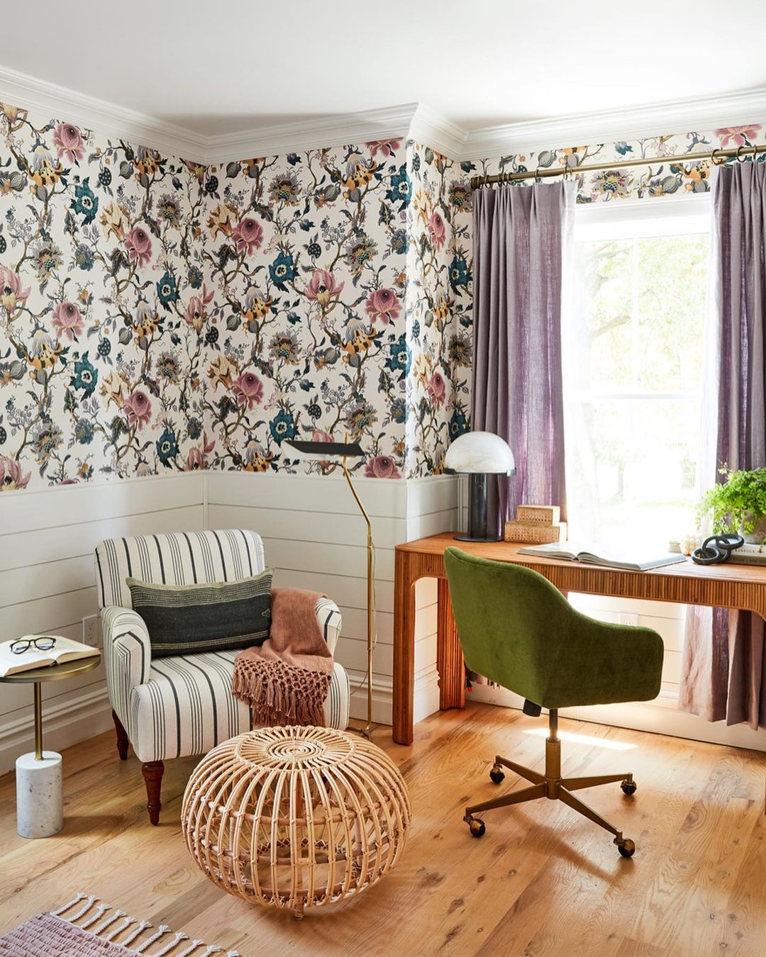 Home office interior with a floral wallpaper, an armchair, a desk, and a green velvet office chair by a window with violet drapes.