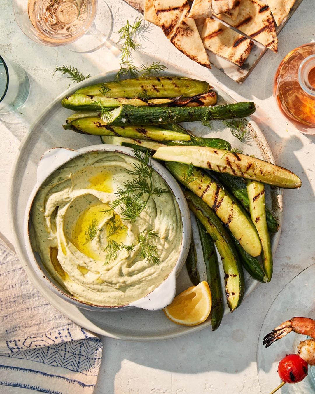 Overhead shot of a plate featuring a bowl of dip, grilled zucchini, pita bread, lemon slice, dill garnish and drinks in the background.