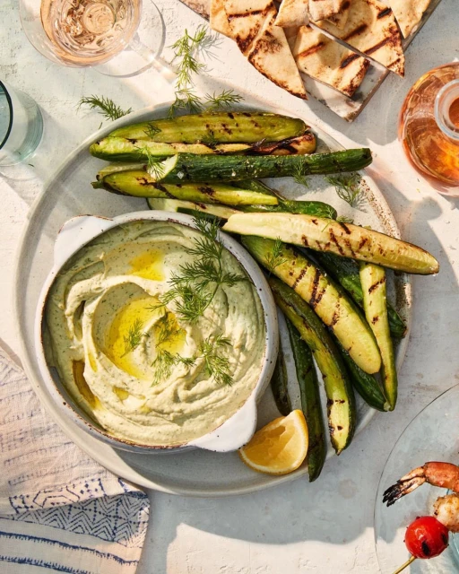 Overhead shot of a plate featuring a bowl of dip, grilled zucchini, pita bread, lemon slice, dill garnish and drinks in the background.