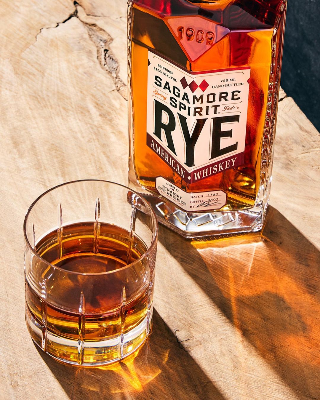 Sagamore Spirit Rye American Whiskey bottle and filled lowball glass on wood surface with dappled sunlight and shadows creating textures.