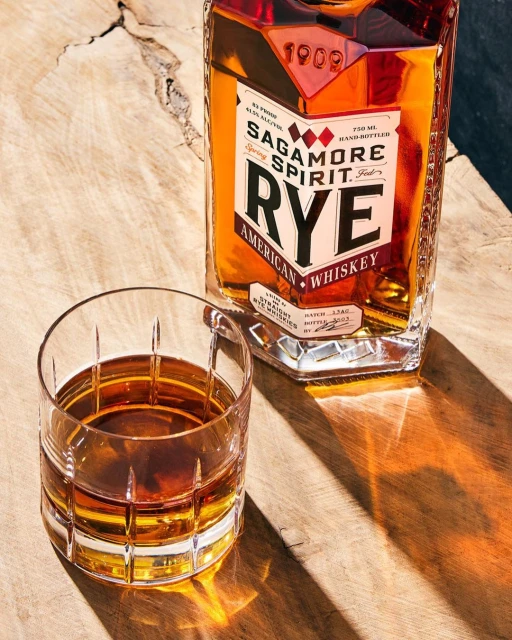 Sagamore Spirit Rye American Whiskey bottle and filled lowball glass on wood surface with dappled sunlight and shadows creating textures.