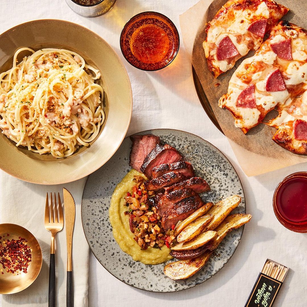 Overhead shot of a steak plate with potatoes and sauce, a plate of spaghetti, and pizza on a tablecloth. Food photography.