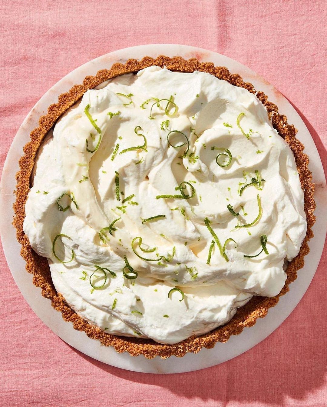 Overhead shot of key lime pie with graham cracker crust and whipped cream topping, decorated with fresh lime zest on pink tablecloth.