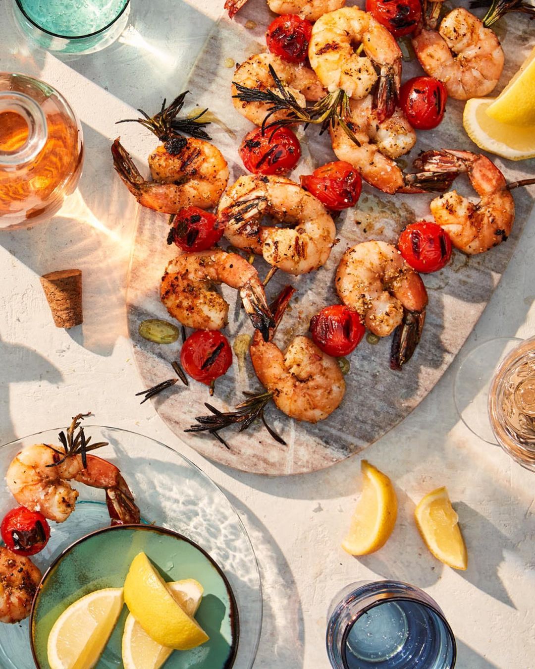 Overhead shot of grilled shrimp skewers with cherry tomatoes, lemon slices, and rosemary sprigs on a marble board and plates.