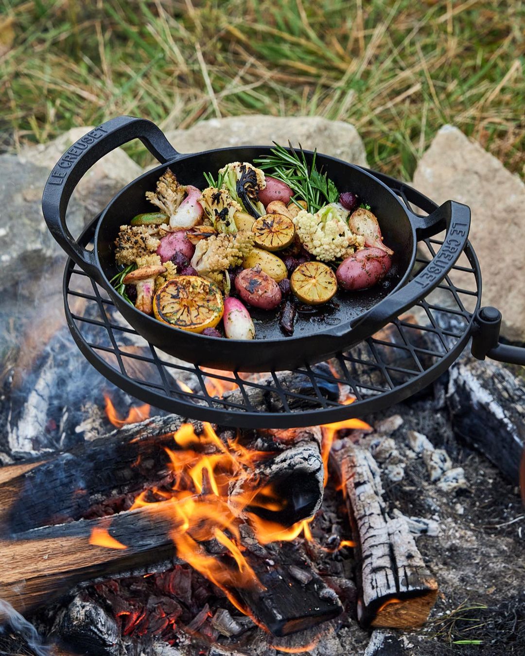 Roasted vegetables with rosemary and lemons in a black cast iron pan over a campfire on a metal grate in an outdoor setting.