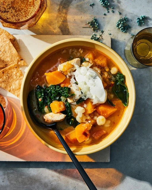 Overhead shot of a yellow bowl filled with chicken tortilla soup garnished with sour cream, alongside drinks and tortilla chips.