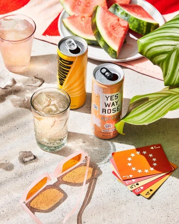 Outdoor summertime still life featuring open beverage cans, cocktails, sunglasses, watermelon slices, and playing cards on a light surface.