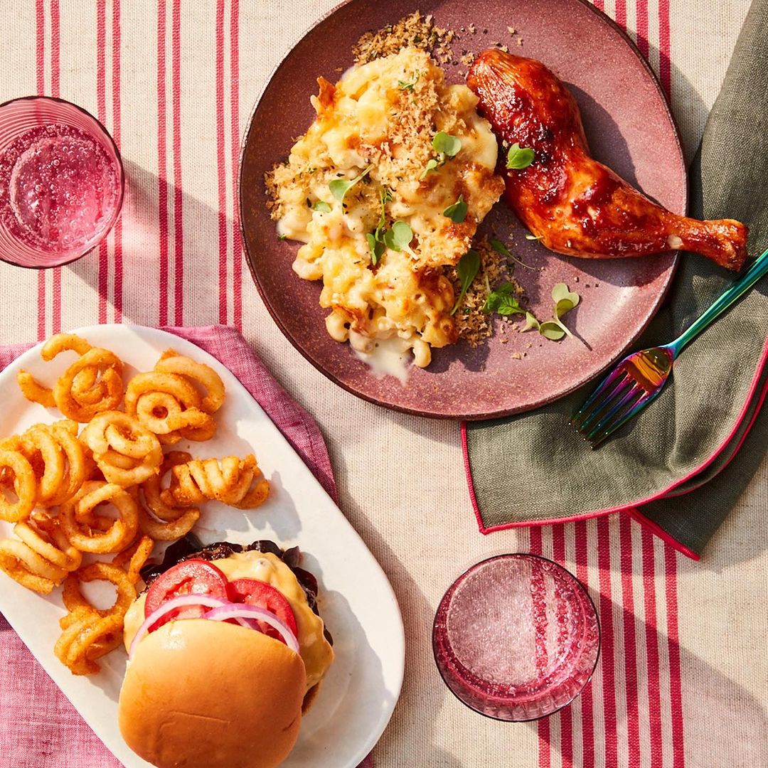 Overhead shot of a table setting with a plate of mac and cheese and chicken, burger and fries, and two drinks, on a striped tablecloth.