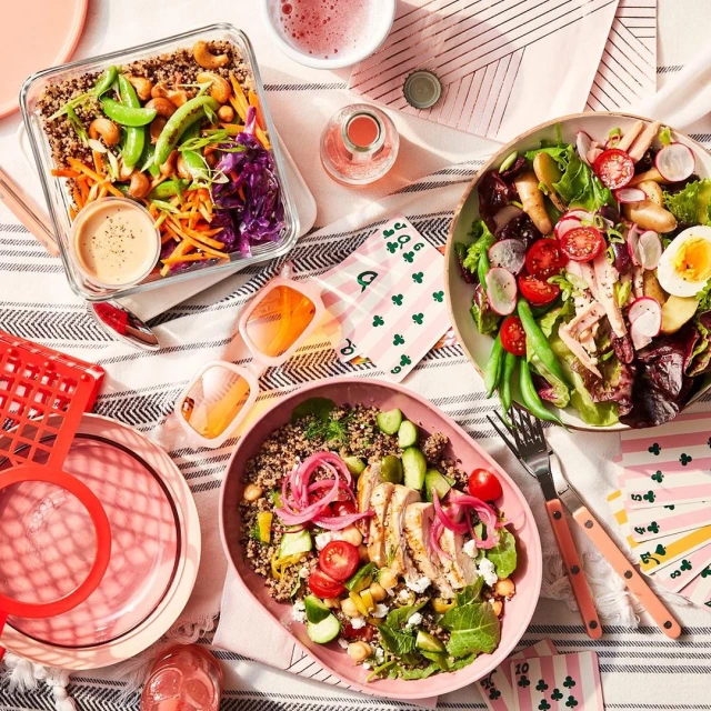 Overhead shot of a vibrant food spread: quinoa and vegetable salads in bowls and containers, complemented by drinks and colorful props.