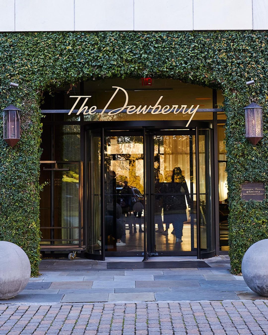 Exterior daytime view of The Dewberry hotel entrance, featuring revolving glass doors, symmetrical greenery, and spherical concrete planters.