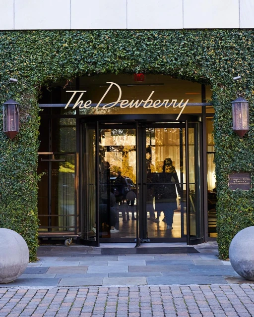 Exterior daytime view of The Dewberry hotel entrance, featuring revolving glass doors, symmetrical greenery, and spherical concrete planters.