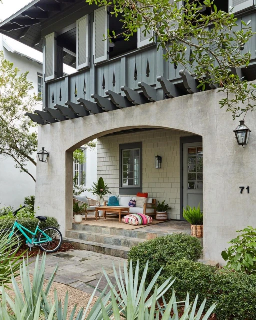 Exterior view of a house with a porch, seating area, and a bicycle parked nearby. Green plants and trees surround the home.