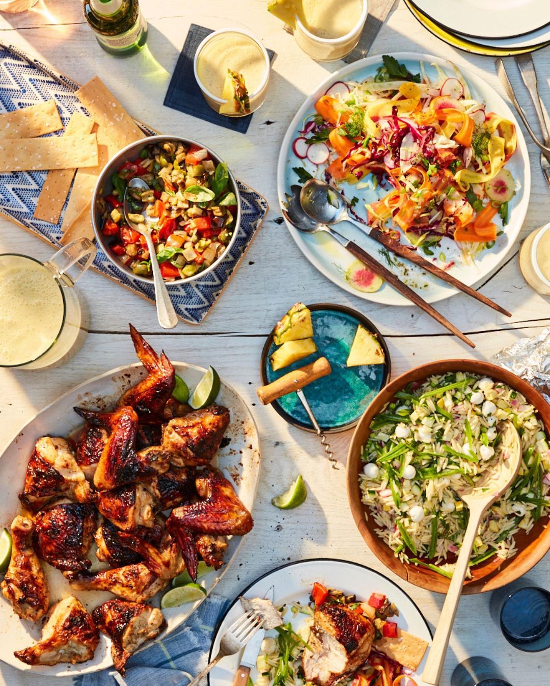 Overhead shot of a buffet spread featuring grilled chicken, salads, rice, drinks, and garnishes arranged on a white wooden table.