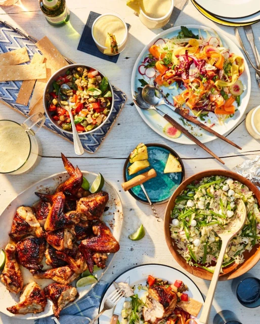 Overhead shot of a buffet spread featuring grilled chicken, salads, rice, drinks, and garnishes arranged on a white wooden table.
