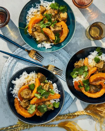 Overhead shot of three bowls of curry with rice, squash, and cilantro, presented on a textured grey surface with metallic forks and drinks.