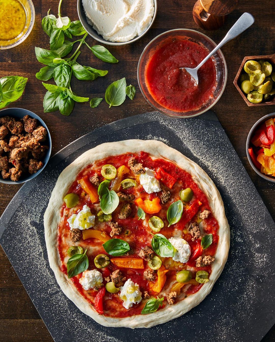 Overhead shot of an uncooked pizza with tomato sauce, mozzarella, basil, peppers, olives, and sausage on a dark wooden table.
