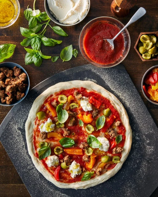 Overhead shot of an uncooked pizza with tomato sauce, mozzarella, basil, peppers, olives, and sausage on a dark wooden table.