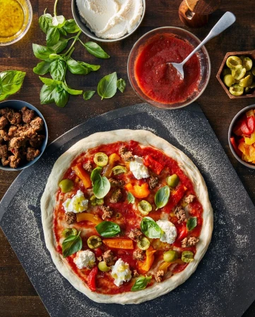 Overhead shot of an uncooked pizza with tomato sauce, mozzarella, basil, peppers, olives, and sausage on a dark wooden table.