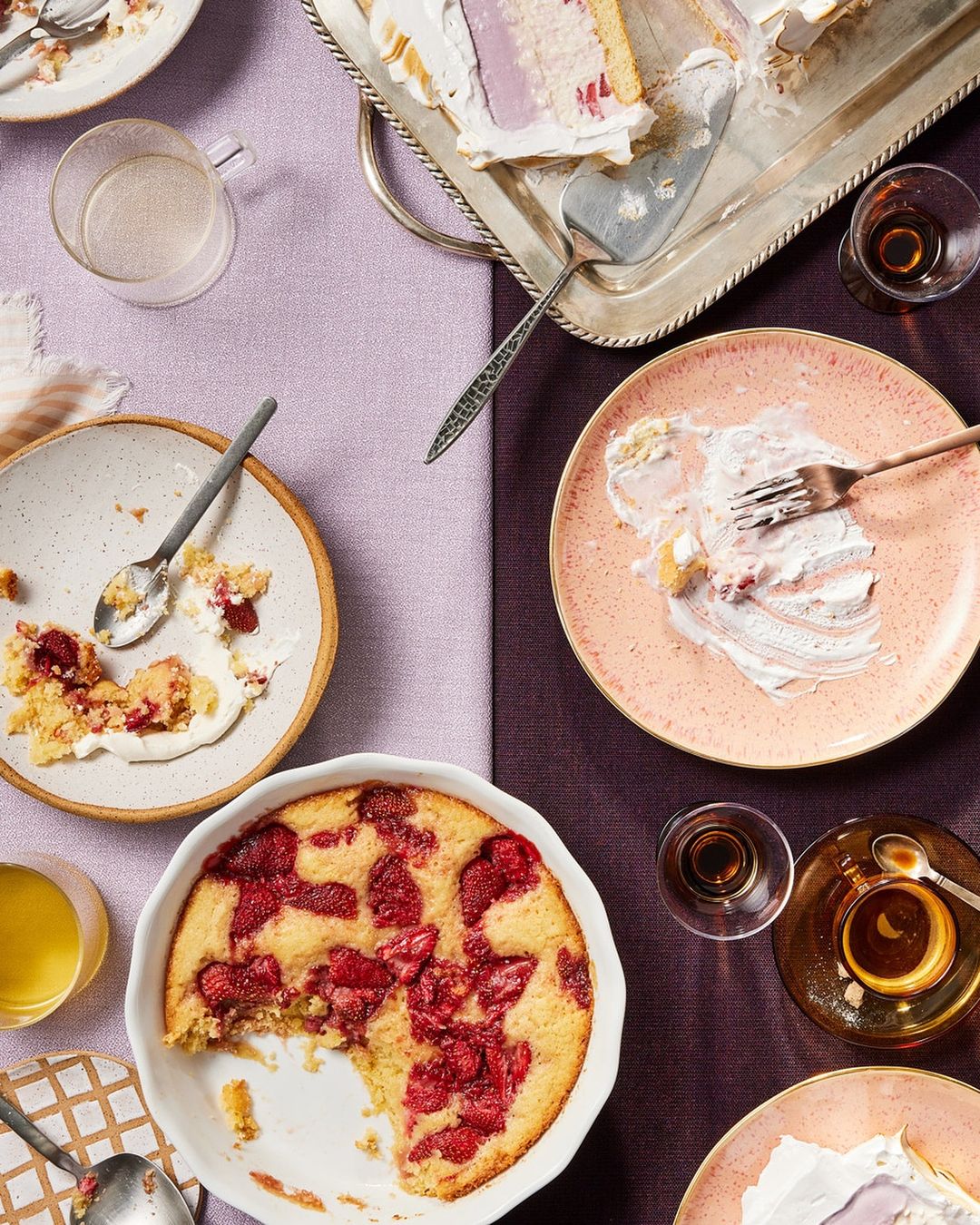 Overhead shot of dessert cakes and pies served on plates and trays, accompanied by glasses of liquor on a two-tone tablecloth.