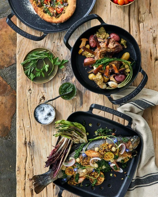 Overhead shot of three black cast iron pans on a wood surface, filled with grilled fish, meat with vegetables, and flatbread.