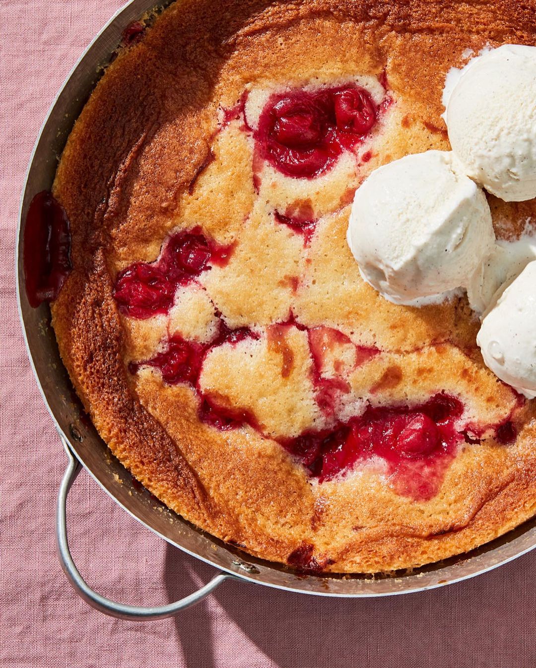 Overhead shot of a baked cobbler dessert with fresh cherries and vanilla ice cream on a pink linen surface in natural lighting.