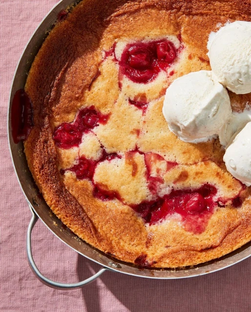Overhead shot of a baked cobbler dessert with fresh cherries and vanilla ice cream on a pink linen surface in natural lighting.