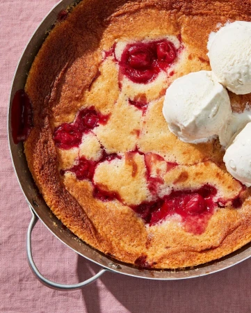 Overhead shot of a baked cobbler dessert with fresh cherries and vanilla ice cream on a pink linen surface in natural lighting.
