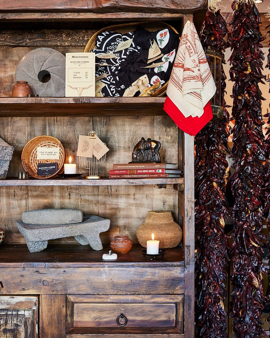 Rustic display on wooden shelves features pottery, woven baskets, books, textiles, chili pepper strands, and vintage decorations.