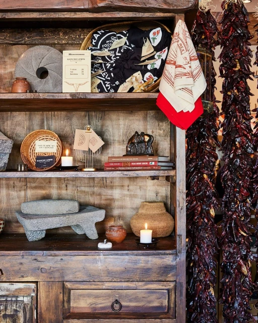 Rustic display on wooden shelves features pottery, woven baskets, books, textiles, chili pepper strands, and vintage decorations.