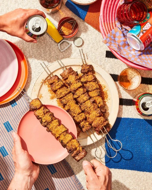 Overhead shot of kebabs on plates held by hands, surrounded by drinks and colorful picnic blanket creating a vibrant culinary scene.