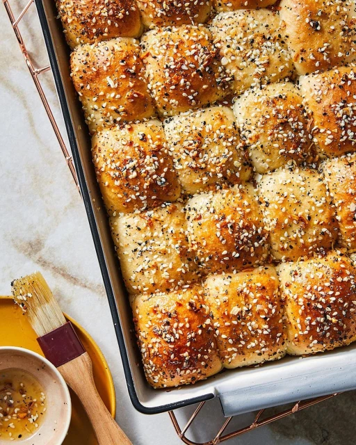 Overhead shot of a tray of golden-brown dinner rolls covered in seasonings, with a small dish of oil and a brush nearby.