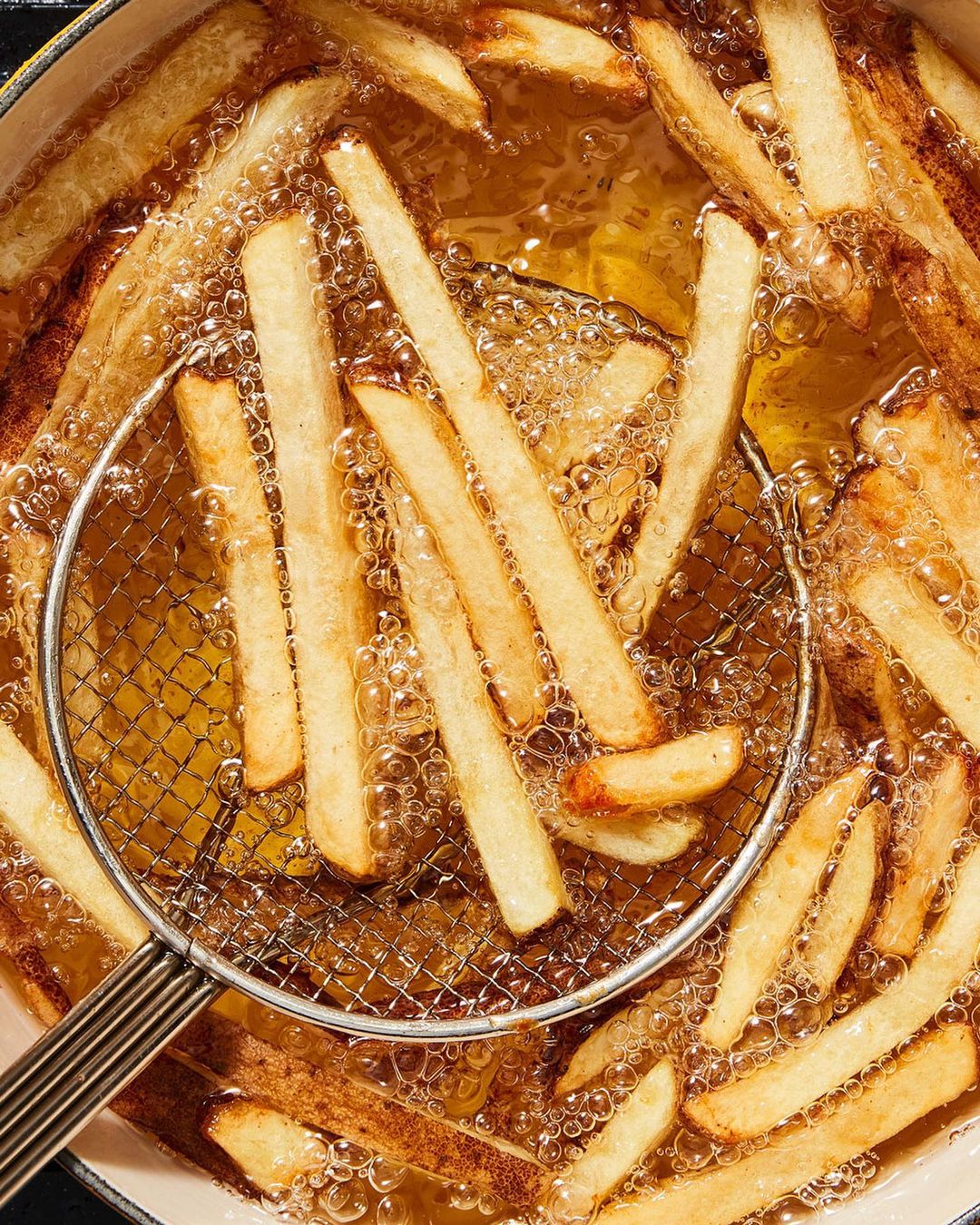 Overhead shot of golden-brown french fries cooking in hot oil, some held in a metal strainer, in a light-colored pot.