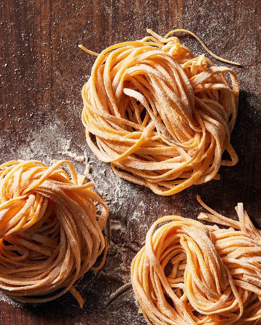 Overhead shot of three nests of dry egg tagliatelle pasta dusted with flour on dark wooden surface, highlighting texture and color.
