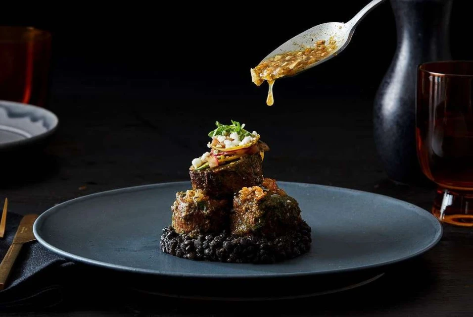 Elevated food presentation of stacked meat rolls on a dark plate with a silver spoon pouring sauce over the dish in a dark setting.