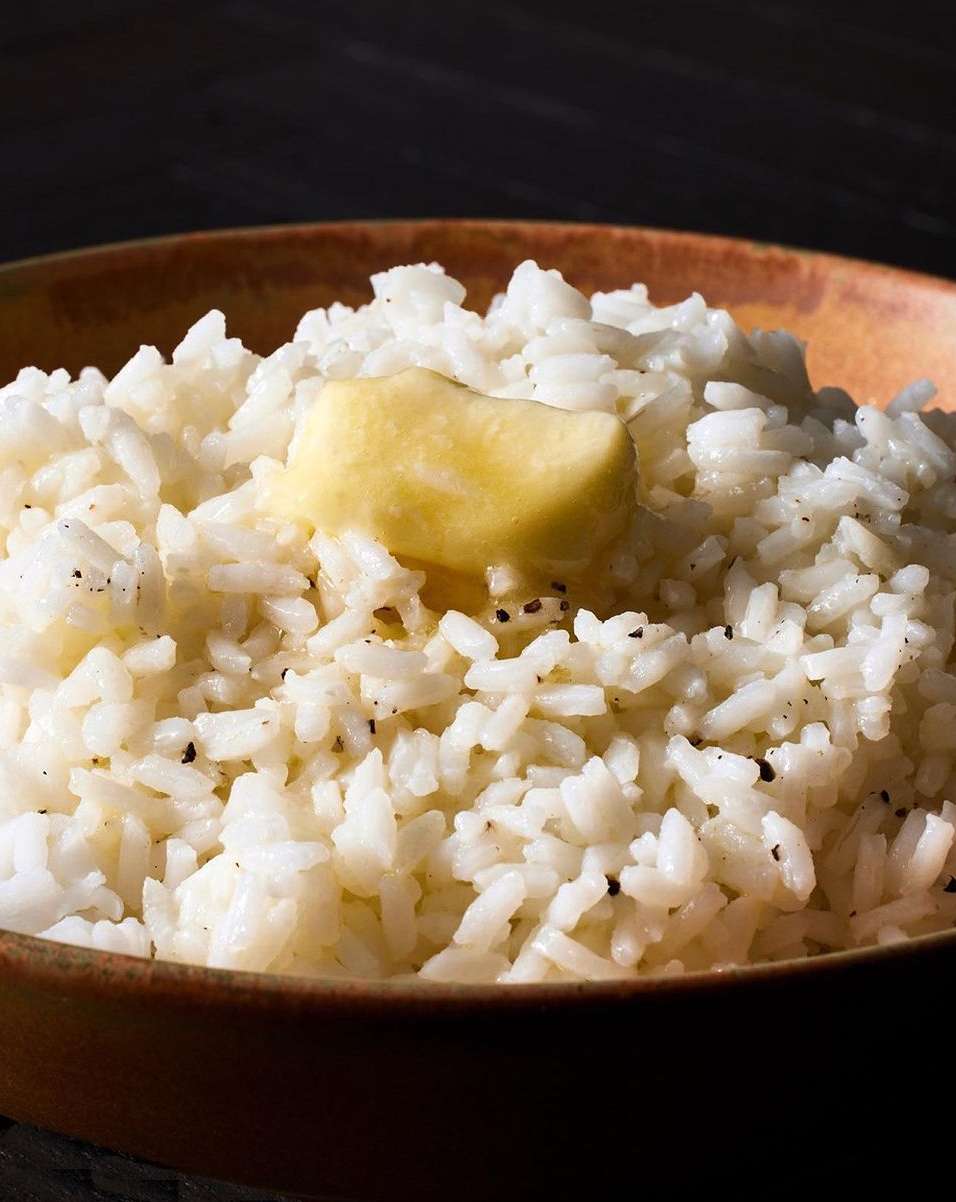 Bowl of white rice with a pat of butter on top, presented against a dark backdrop, showcasing a simple, comforting food scene.