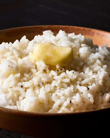 Bowl of white rice with a pat of butter on top, presented against a dark backdrop, showcasing a simple, comforting food scene.