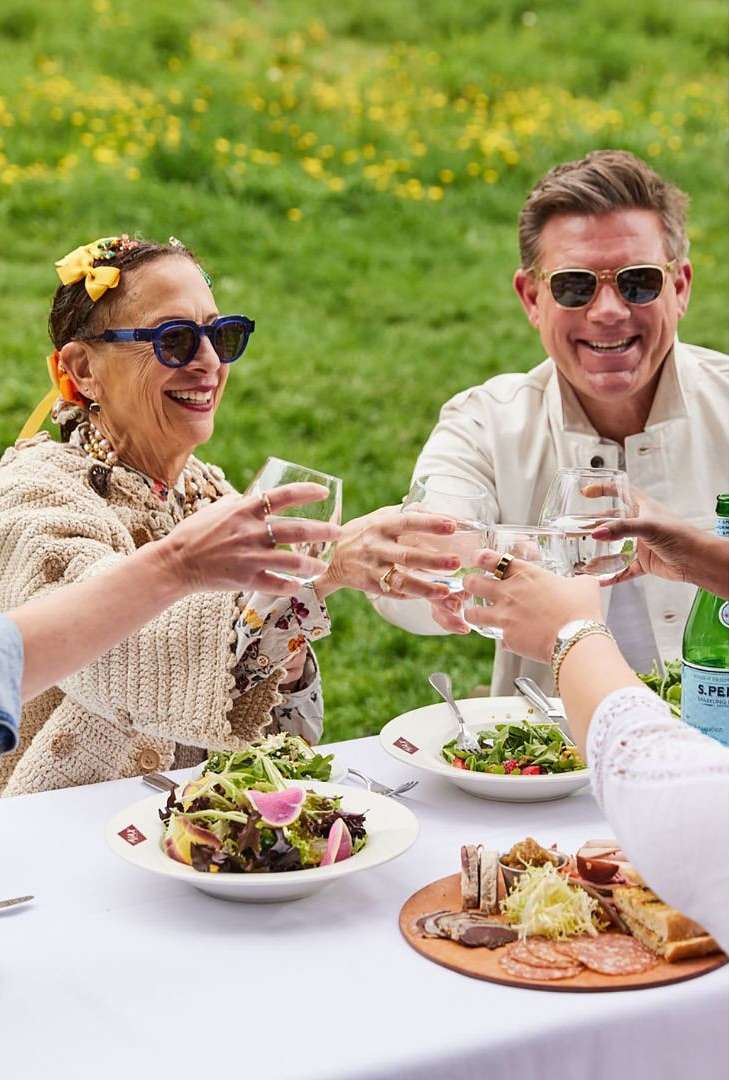 Group of people toasting with wine glasses around a table with salads and charcuterie outdoors on a sunny day in a grassy field.