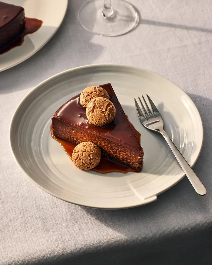A slice of chocolate cake with caramel glaze and small cookies on a ceramic plate with a fork, photographed by Christopher Testani