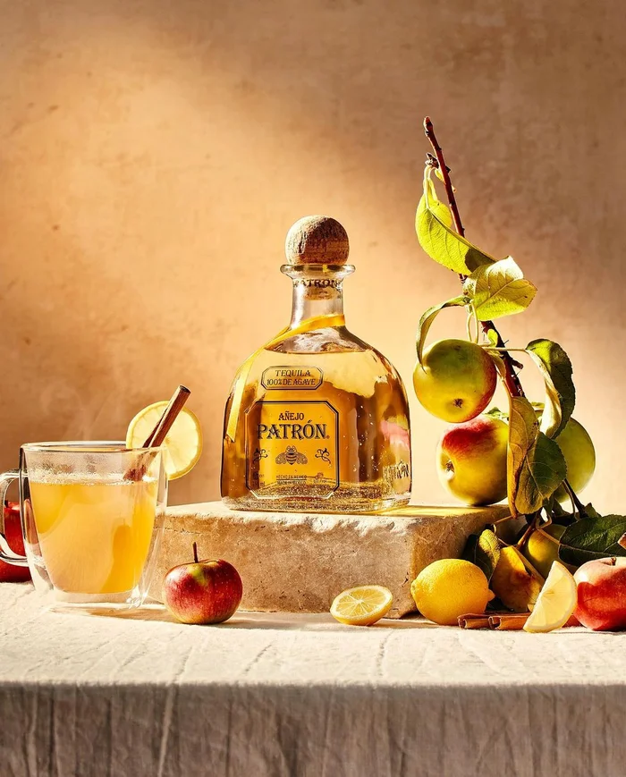 Patron Anejo tequila bottle on stone block with apples, lemons, and a cocktail glass by Christopher Testani