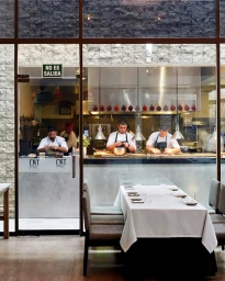 Chefs working in a restaurant kitchen viewed through a large glass window behind a set dining table by Christopher Testani